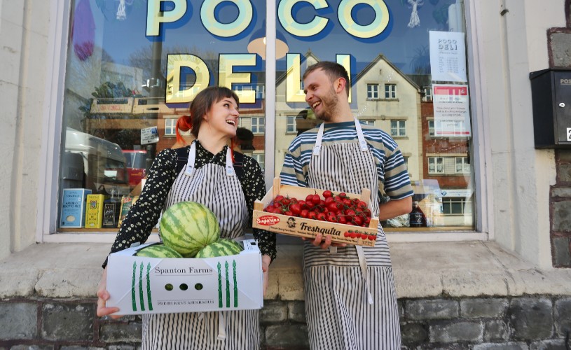 Two people holding boxes of fruit and veg in front of Poco Deli